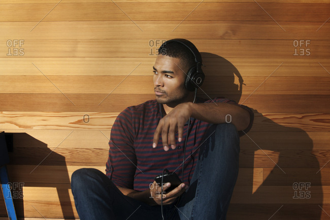A young man leans against a wood paneled wall and wears headphones