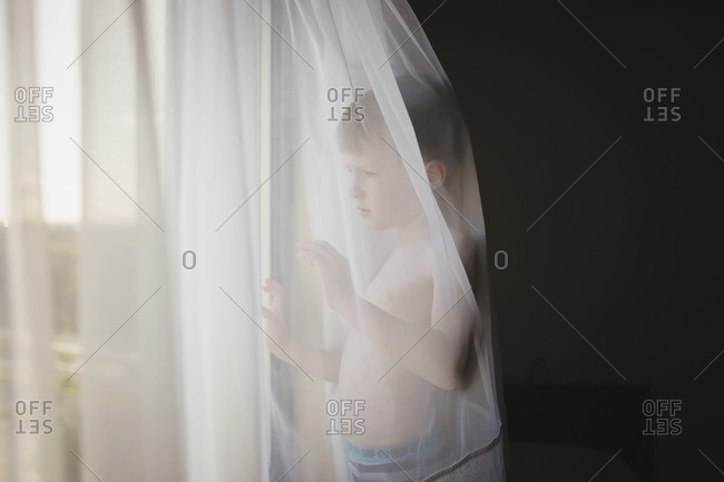 Boy with no shirt stands behind curtain looking out window