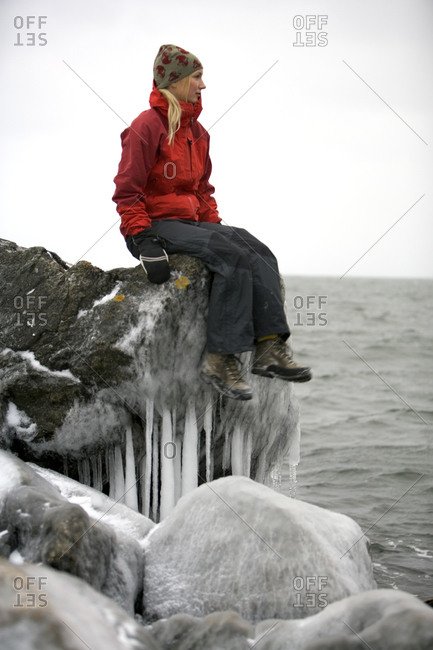 A woman sits on a rock covered in ice and looks at the ocean