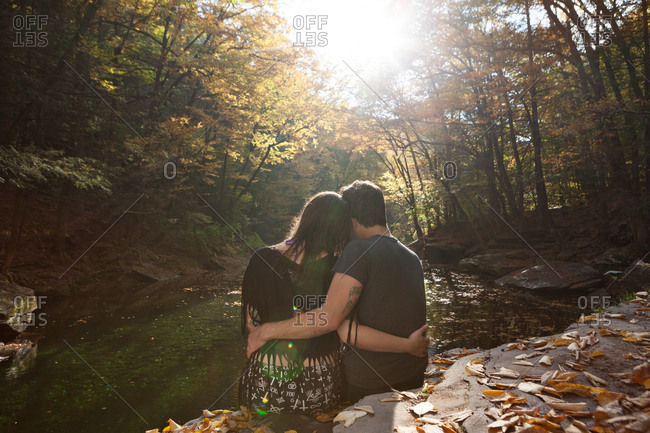 Romantic young couple seated with arms around each other on a rock above a stream