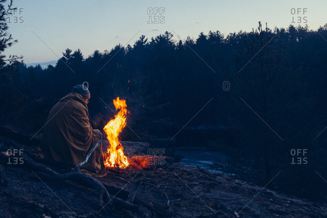 Man wrapped in a blanket sits at a campfire