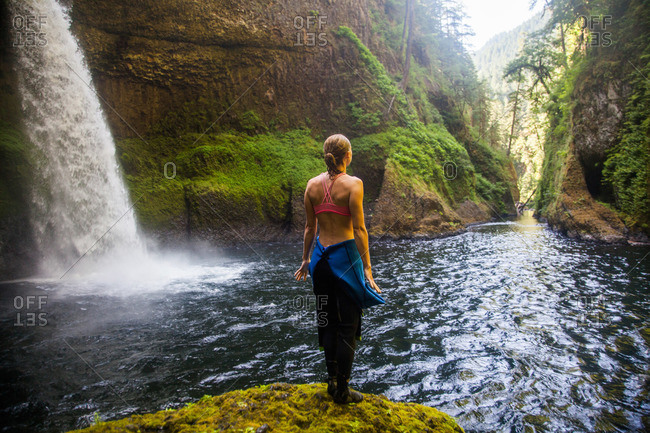 A woman stands on the edge of a rocky outcrop in a wetsuit overlooking Eagle Creek Falls in the Columbia Gorge, Oregon