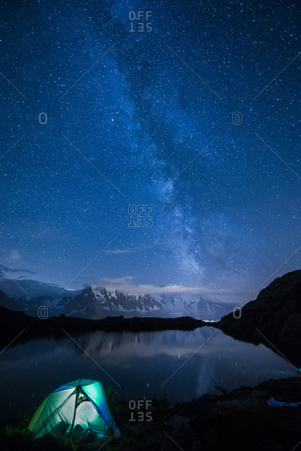 Lit tent on the shore of the lake by night with Milky way and Mount Blanc reflected in the lake
