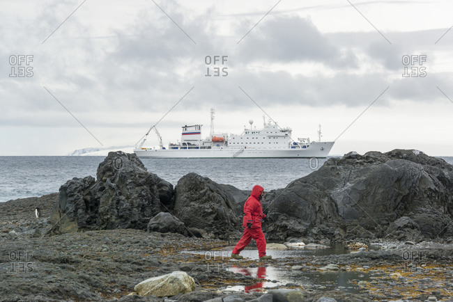 Man walking along rocks by the sea in the Antarctic with a polar research vessel in the background