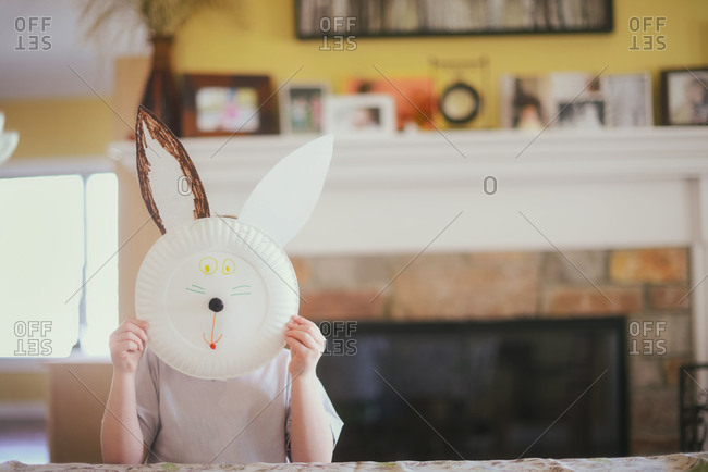 Young child holds up rabbit face made of paper plate in living room