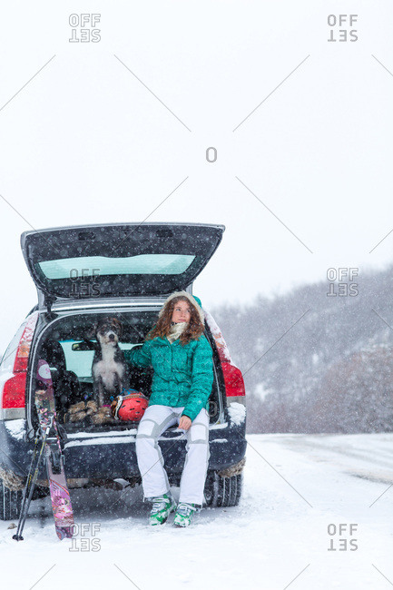 Young woman and her dog sit in their vehicle hatch with skis in snowstorm