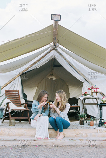Two women sitting outside a large tent laughing and having a glass of wine