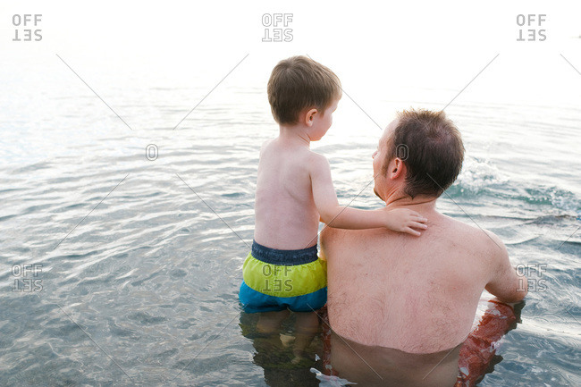 A father sits in calm, shallow water with his son