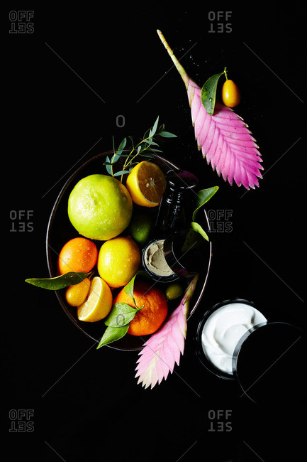 Natural face cream and face mask in black ceramic bowl with citrus fruits and tropical ginger flowers on black background