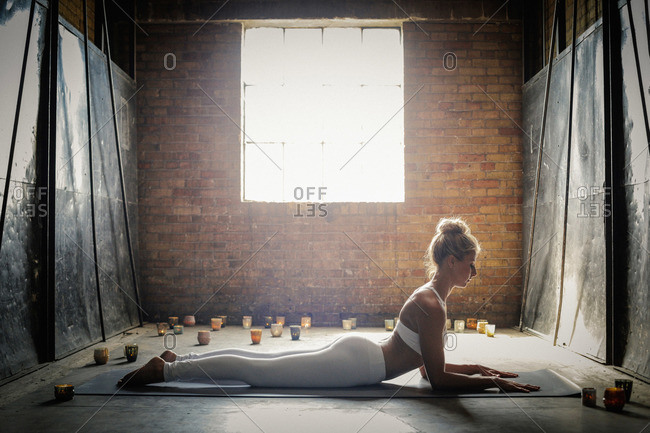 A blonde woman, in a white crop top and leggings, lying on a yoga mat on the floor surrounded by candles, doing yoga