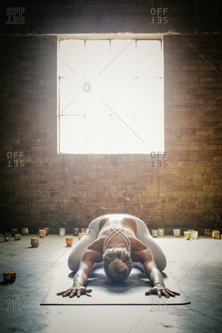 A blonde woman in a white crop top and leggings, bending down on a yoga mat surrounded by candles, doing yoga