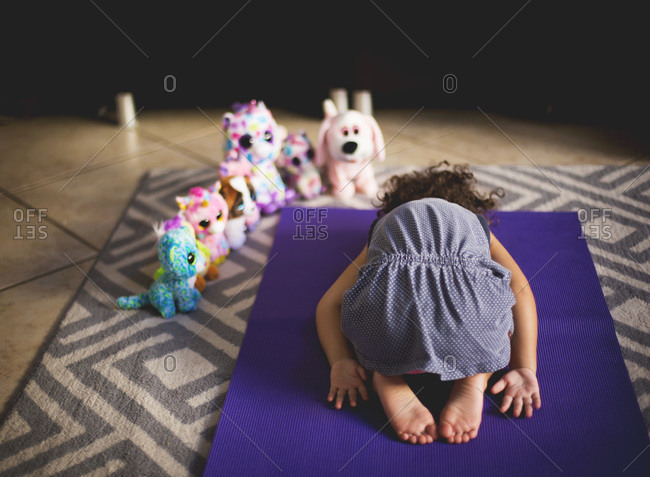 A little girl bends over on a yoga on a mat surrounded by stuffed animals