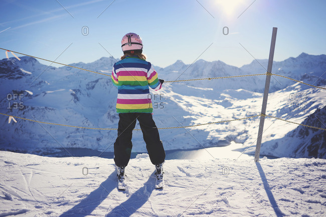 Young girl in striped ski jacket holds onto rope fence while overlooking mountain landscape