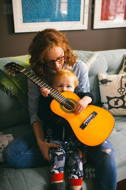 Woman holds her toddler son on her lap as he plays on toy guitar