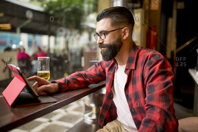 Adult bearded hipster style man touching tablet in bar and drinking alcohol near to window