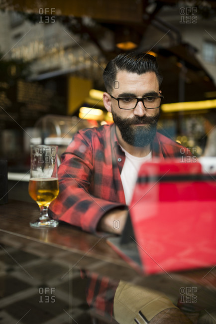 Adult bearded hipster style man touching tablet in bar and drinking alcohol near to window