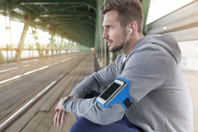 Young athletic man with earbuds and wearable computer sitting on bridge