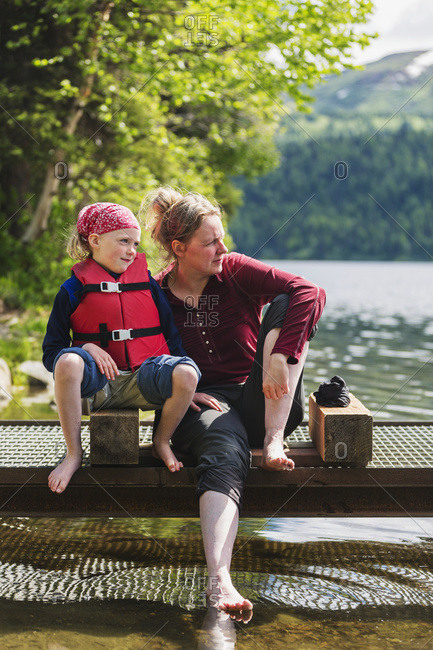 A mother and daughter sits on a dock looking out over Byers Lake, soaking their bare feet in the water, Denali State Park, Alaska, United States of America