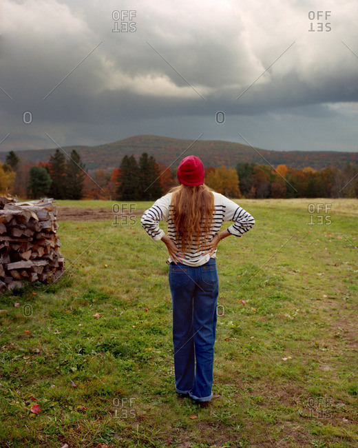 Back view of woman with hands on her hips standing in field next to wood pile in fall