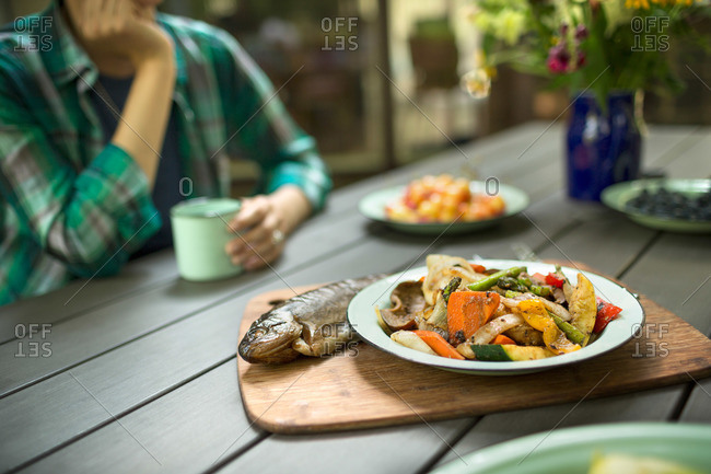 A person seated at a table outdoors A cooked fresh fish and dish of vegetables