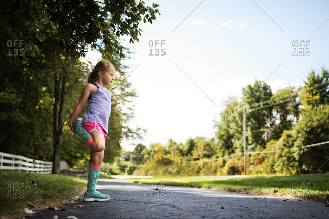 Young girl in knee socks and running shorts doing leg stretches on country road