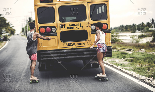 Two teens riding skateboards while holding onto the back of a school bus