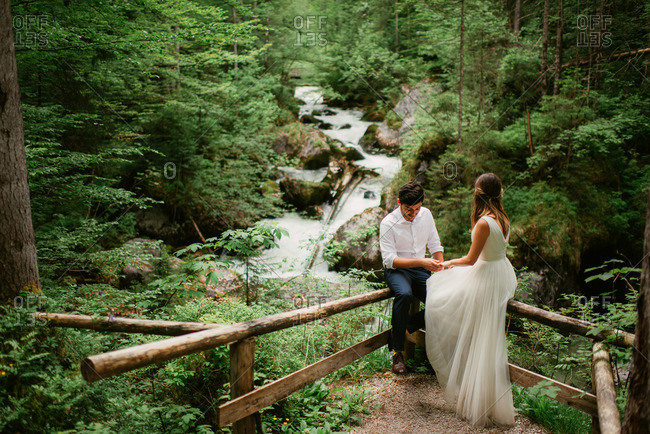 Newlyweds holding hands as they sit on a wooden fence by a river