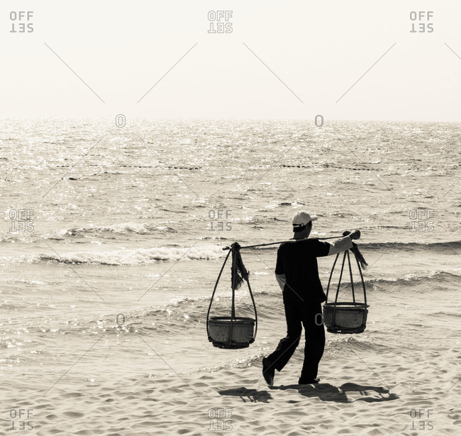A beach vendor carrying his produce using a shoulder pole or \'Yoke\' along a beach in Phu Quoc Island, Vietnam