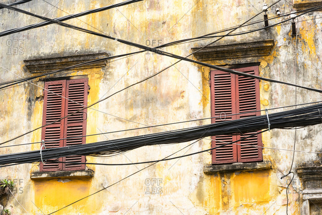 An old yellow building with two windows with brown shutters with a chaotic collection of cables in the foreground in the city of Hanoi, Vietnam