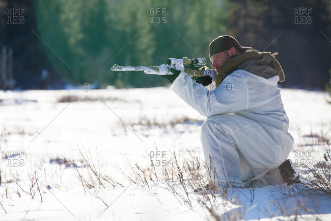 A man wearing a cold-weather camouflage outfit aims a rifle while hunting in a snow-covered field