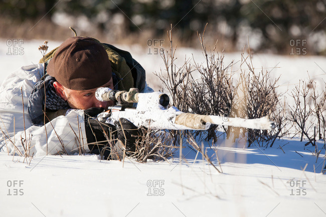 A hunter wearing a cold-weather camouflage outfit aims a rifle at his target while laying down in a snow-covered field