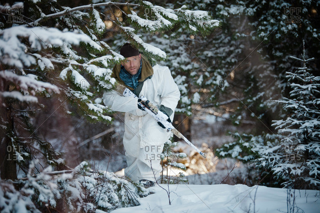 A man wearing a cold-weather camouflage outfit carefully moves through the forest towards his target