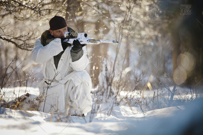 A hunter wearing a cold-weather camouflage outfit aims a rifle while crouching in a snow-covered forest