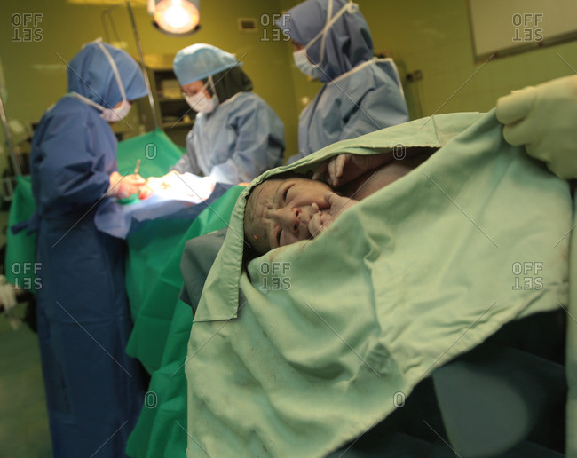 Newborn baby in foreground with medical team completing a C-section