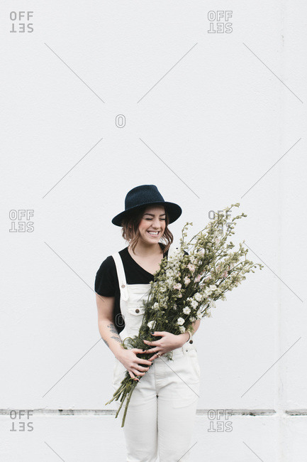 Laughing woman in black hat with white overalls holds white flowers against white wall