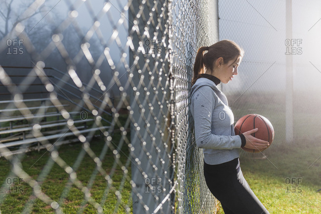 Young woman with basketball leaning against fence on a sports field