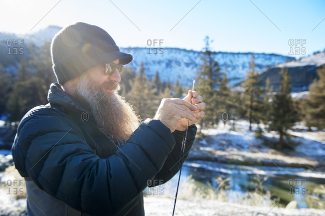 Bearded fisherman threading fishing line through rod by river in winter