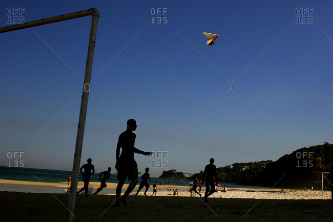 A hang glider in the late afternoon light above football players at Sao Conrado beach