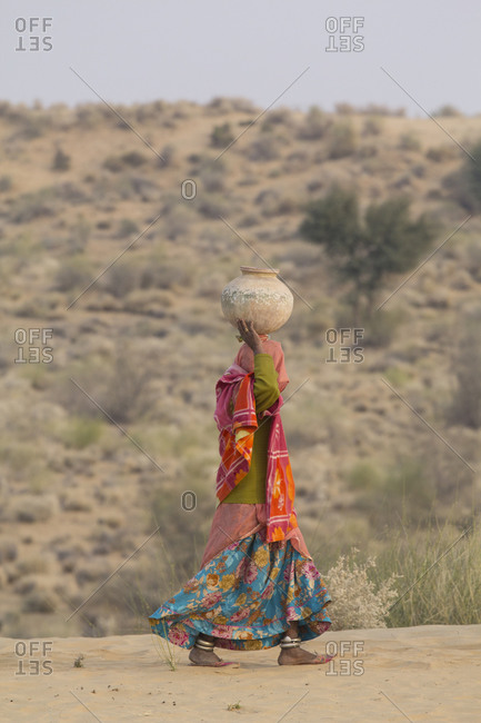 Colorfully dressed village woman walking with pot on her head, going to village well