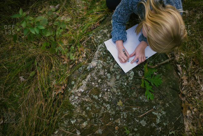 Overhead view of young girl writing with crayons on paper on rock in woods