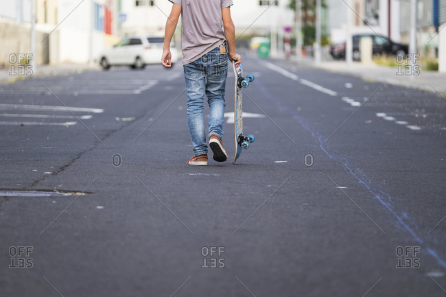 Back view of boy carrying his skateboard on a street