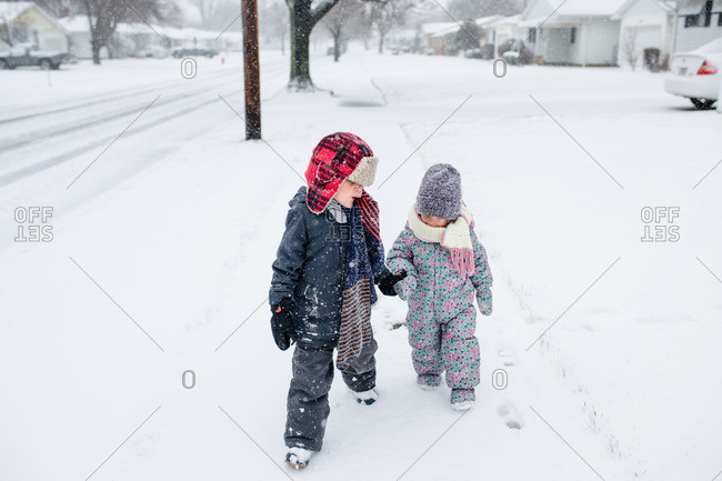 Boy holding his toddler sister\'s hand as they walk together on sidewalk during snowstorm