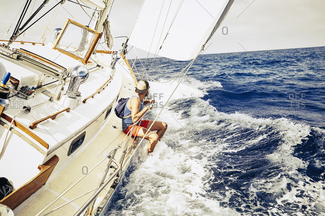 Young man sitting on the side of a sailboat with his legs in the water
