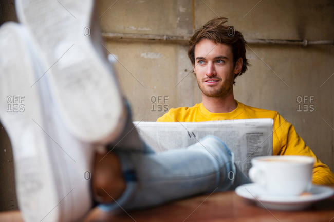 Young man sitting in cafe with feet on table and reading newspaper