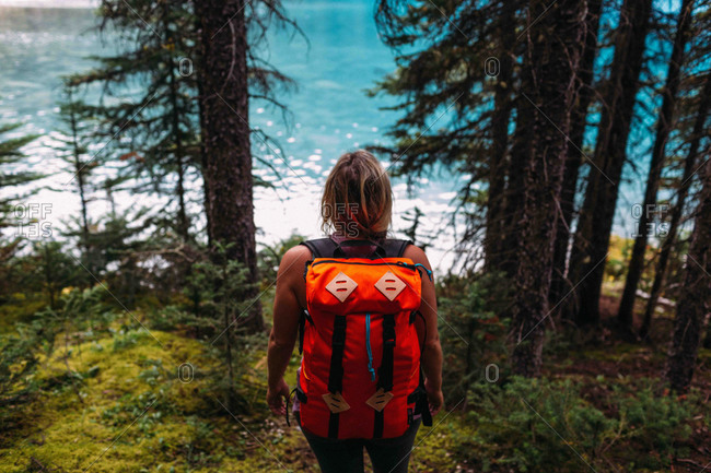 High angle rear view of mid adult woman carrying orange color backpack standing in forest looking at water, Moraine lake, Banff National Park, Alberta Canada