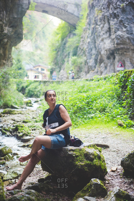 Woman sitting on rock in stream, rock hills and stone bridge in background, Garda, Italy