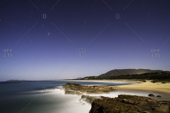 Stars streaking through a night sky on the east coast of Australia Norther brother in the background lit up by the town of Laurieton