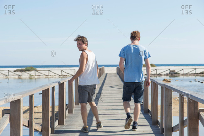 Full length rear view of young men walking on wooden pier, looking over shoulder, Sardinia, Italy