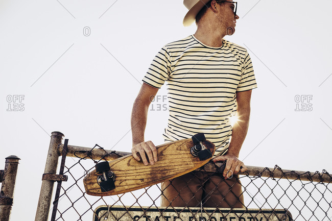 Man in hat and sunglasses climbing over fence with skateboard