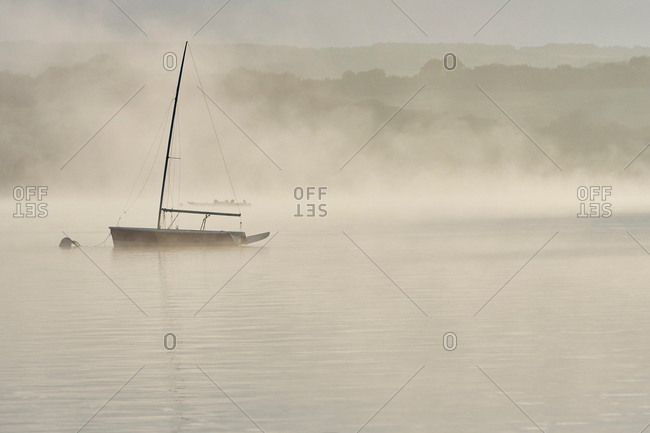 Silhouetted yacht on misty Wimbleball lake at dawn, Exmoor, Somerset, England